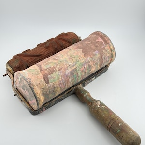 May include: A vintage dual-roller paint tool featuring a textured brown rubber patterned roller and a smooth cylindrical roller covered in layers of dried, multi-colored paint. The metal frame and wooden handle show significant wear and paint residue.