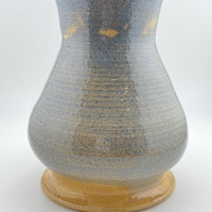 May include: A ceramic vase with a flared rim and a rounded body, featuring a speckled blue and grey glaze with horizontal ridges throughout. The base of the vase is a solid, unglazed tan color, providing a distinct contrast to the upper section.