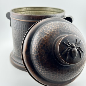 May include: A cylindrical copper-toned container with a hammered texture and two side handles. The matching lid features a raised, three-dimensional spider figure centered on the top. The interior of the container is metallic and smooth.