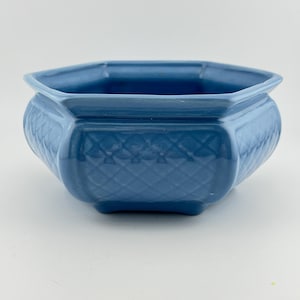 May include: A hexagonal ceramic planter in a solid medium blue glaze. The exterior features a raised diamond lattice pattern across each of the six sides. The vessel has a wide, flared rim and sits on a small, integrated base with four feet.