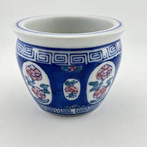 May include: A small ceramic bowl featuring a white base with a deep blue background. The exterior is decorated with repeating geometric patterns near the rim and base, interspersed with oval panels containing pink floral motifs and green leaves.