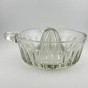 May include: A clear glass citrus juicer featuring a central reamer cone and a surrounding bowl with a textured, vertical ribbed pattern. The juicer includes a small, integrated handle on one side and a pouring spout on the opposite side.