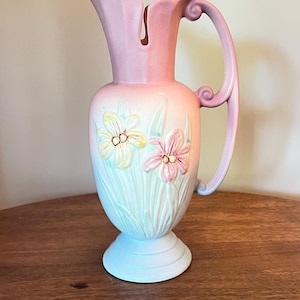 May include: A pastel-colored pitcher with a floral design. The pitcher features a light pink handle and spout, with a gradient of colors from light blue to pink. Two embossed flowers are on the front.