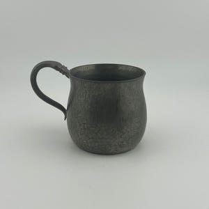 May include: A small, antique pewter mug with a rounded body and a curved handle. The mug has a dark, textured surface with a slightly cracked appearance. The handle is attached to the side of the mug, and the interior is dark.