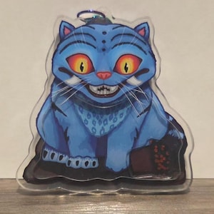Peut inclure: Un charm en acrylique en forme de chat bleu avec un sourire menaçant et de grands yeux jaunes. Le chat a un corps bleu avec des rayures bleu foncé et un collier à motifs. Le charm a une boucle argentée en haut pour l'accrocher.