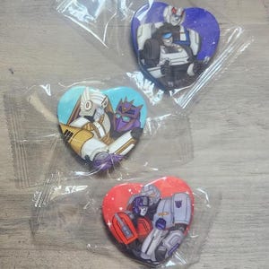 May include: Three heart-shaped pins featuring Transformers characters. The first pin is purple with a white and blue robot. The second pin is yellow with a gold and purple robot. The third pin is red with a red and gray robot.