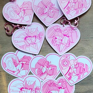 May include: A selection of heart-shaped keyrings with pink and white robot illustrations. Each keyring has a rose gold-coloured clasp. The robots are shown in a romantic pose. These keyrings are a collectible item.