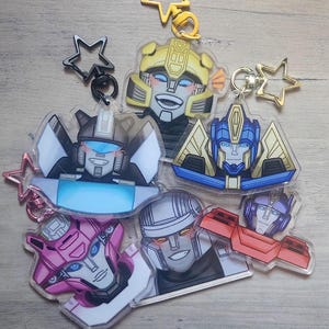Op de afbeelding: Set van 7 Transformers-personage sleutelhangers. De sleutelhangers zijn gemaakt van helder acryl met kleurrijke cartoonafbeeldingen van de personages. De personages omvatten Bumblebee, Optimus Prime, Starscream, Megatron, Arcee en Bulkhead.