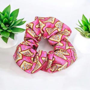 May include: A pink scrunchie featuring a repeating pattern of toast slices with colourful sprinkles. The fabric is gathered, giving a voluminous, textured look. This is a hair accessory.