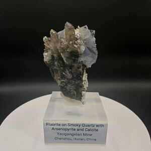 May include: A natural mineral specimen featuring fluorite on smoky quartz with arsenopyrite and calcite. The crystal cluster displays a mix of clear, brown, and blue hues, with a dark green base. The specimen is mounted on a clear acrylic base with a label.