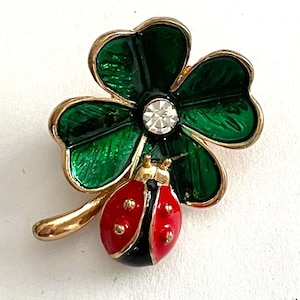 May include: A gold-toned brooch featuring a green four-leaf clover with a central clear rhinestone. A red and black ladybug with gold accents rests on a gold stem, adding a touch of whimsy to this jewelry piece.