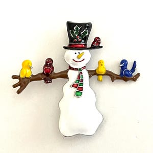 May include: A whimsical snowman brooch with a black top hat adorned with holly and berries. The snowman has a red and green scarf and is perched on a brown branch with colorful birds. The brooch is made of enamel.