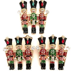 May include: Three gold-tone metal brooches, each featuring a trio of nutcracker figures. The figures are decorated with red, green, and black enamel, accented with clear and colored rhinestones on their hats, uniforms, and instruments.