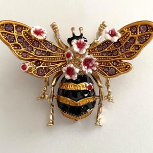 May include: A gold-toned bee brooch with intricate detailing. The wings are adorned with purple rhinestones and outlined in gold. The bee's body is black and gold striped, with small red and white flower accents. A decorative accessory.