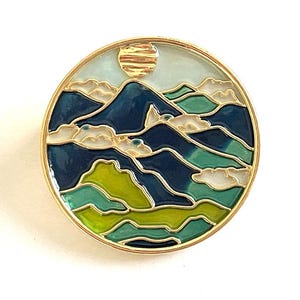 May include: A gold-toned round pin with a blue, green, and yellow enamel design of mountains and clouds. The sun is depicted in the top center of the pin.