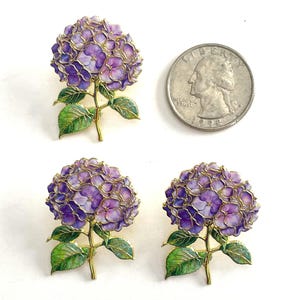 May include: Three gold-toned metal hydrangea flower brooches with purple and blue enamel petals and green enamel leaves. A US quarter coin is placed next to the top brooch for scale, showing the flower head is approximately 0.95 inches wide.