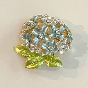 May include: A floral brooch featuring a cluster of light blue and white flowers with sparkling crystal accents. The brooch has gold-tone metal detailing and green enamel leaves. A decorative accessory.