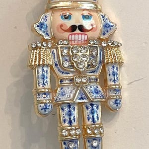 May include: A gold-tone nutcracker brooch featuring white enamel with blue floral patterns, accented by clear rhinestones on the crown, shoulders, chest, and boots. The figure has a painted face with blue eyes, a brown mustache, and pink lips.