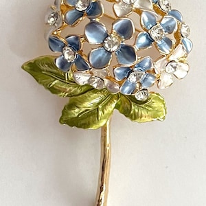 May include: A floral brooch in the shape of a hydrangea. The brooch features blue and white enamel flowers with rhinestone accents. The stem and leaves are gold-toned metal with green enamel.
