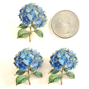 May include: Three blue hydrangea flower brooches with green leaves and gold stems. The brooches are next to a quarter for size comparison. The flowers have varying shades of blue and are detailed with gold outlines.