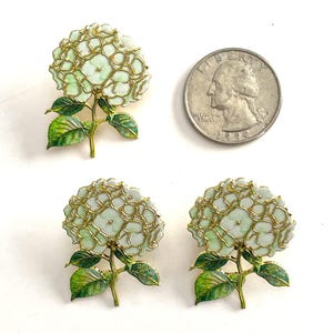 May include: Three gold-toned metal hydrangea flower pins with pale green and white enamel petals and green leaves, shown next to a United States quarter coin for scale. Each pin features a detailed floral cluster with a stem and two leaves.