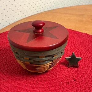 May include: A round woven basket with a dark red wooden lid featuring a star design and a central knob. The basket body has alternating bands of natural tan and muted gray woven material. A small, dark gray metal star ornament sits beside the basket.