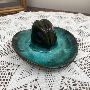 May include: A green and brown ceramic cowboy hat shaped trinket dish. The dish is on a white crocheted doily.