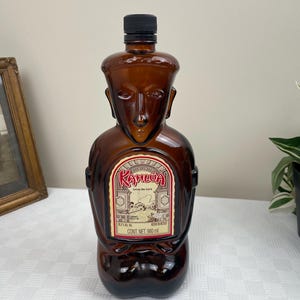 May include: A brown glass bottle of Kahlua liqueur shaped like a person. The bottle has a label with the text "Kahlua" and "Liqueur de Cafe". The bottle is 980 ml.
