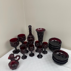 May include: A set of ruby red glass tableware, including a decanter, a jug, a vase, bowls, and wine glasses. The glassware has a textured, diamond-like pattern.