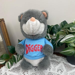 May include: A grey plush bear wearing a light blue shirt with the word "DIGGER" in red letters. The bear has a white face and pink nose and ears. The bear is sitting on a white lace tablecloth.
