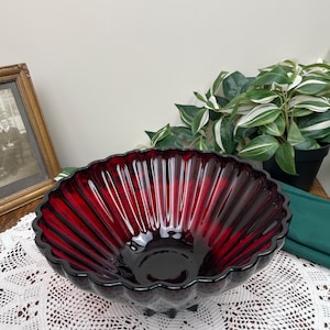May include: A large, deep red glass bowl with a fluted design and a scalloped rim. The bowl is sitting on a white crocheted doily.
