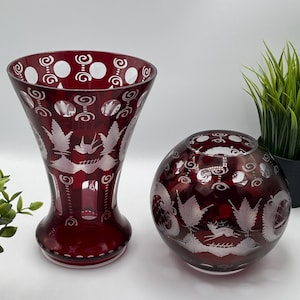 May include: Two ruby red glass vases. One is a tall, flared vase with an etched design of birds and foliage. The other is a round, spherical vase with a similar etched design. Both vases have a decorative, vintage aesthetic.