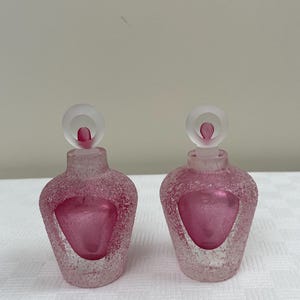May include: Two frosted glass perfume bottles with pink glass stoppers and a textured surface.