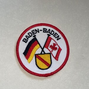 Peut inclure: Écusson brodé circulaire avec bordure rouge et fond blanc. Le texte "BADEN-BADEN" en noir est présent, avec les drapeaux croisés de l'Allemagne et du Canada. Un écusson jaune avec une bande diagonale rouge est en dessous.
