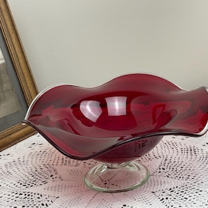 May include: A ruby red glass bowl with a wavy rim and a clear glass pedestal base. The bowl is sitting on a white crocheted tablecloth.