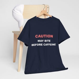 Puede incluir: Camiseta azul marino con el texto "CAUTION MAY BITE BEFORE CAFFEINE" en letras blancas y coral. Una gorra de béisbol blanca descansa sobre una silla de madera detrás de la camiseta.