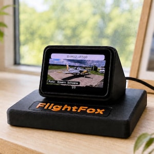 Op de afbeelding: Een zwart digitaal weergaveapparaat met de tekst FlightFox in oranje op de basis. Het scherm toont een afbeelding van een klein vliegtuig op een landingsbaan met de tekst D-MIIZ-P300 en Foto: Bjoern Schoene. Het apparaat is verbonden met een zwarte kabel.