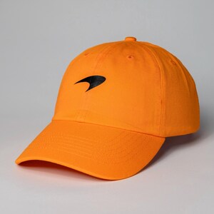 Könnte beinhalten: Eine leuchtend orangefarbene Baseballkappe mit einem schwarzen Logo auf der Vorderseite. Das Logo ist ein stilisierter Schwung.