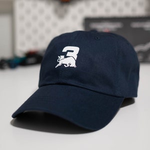 Puede incluir: Gorra de béisbol azul marino con un logotipo blanco que muestra el número 3 y un toro. La gorra tiene una visera curva y un diseño clásico de seis paneles. Un accesorio elegante para uso casual.
