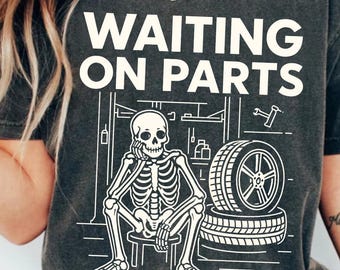 May include: Dark gray t-shirt with the text "WAITING ON PARTS" in white. The graphic shows a skeleton sitting on a stool, leaning on its hand, with tires and tools in the background. The shirt is a casual wear item.