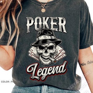 Puede incluir: Camiseta gris oscuro con un diseño gráfico de una calavera con sombrero y fumando un cigarro. La palabra "POKER" está encima de la calavera y "Legend" debajo, en fuente roja y blanca. Tema de póquer.