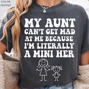 May include: Dark gray t-shirt with the text "MY AUNT CAN'T GET MAD AT ME BECAUSE I'M LITERALLY A MINI HER" in white, along with a stick figure illustration of an adult and a child. The shirt is a pepper color.