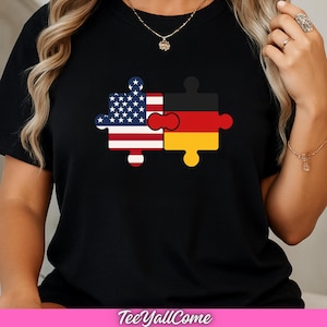 Op de afbeelding: Zwart T-shirt met een puzzelstuk ontwerp. Eén stuk toont de Amerikaanse vlag, de andere de Duitse vlag. Het shirt heeft de tekst "TeeYallCome" onderaan.
