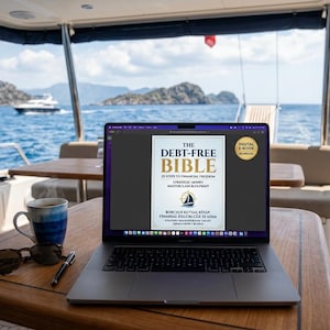 May include: A laptop displaying "The Debt-Free Bible" sits on a wooden table aboard a yacht. Beside it are sunglasses, a pen, and a blue and white mug. The background shows a seascape with a boat and islands.