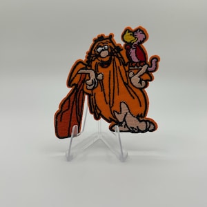 Peut inclure: Un écusson en tissu brodé représentant un personnage de dessin animé aux longs cheveux et à la barbe orange, vêtu d'un vêtement orange, tenant une petite créature rose et rouge sur une main et un grand objet en forme de pagaie orange.