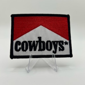Könnte beinhalten: Ein rechteckiger bestickter Aufnäher mit einem roten oberen Bereich und einem weißen unteren Bereich mit schwarzem Rand. Das Wort cowboys ist in schwarzen Kleinbuchstaben in der Mitte aufgestickt, mit einem kleinen schwarzen Stern neben dem Text.