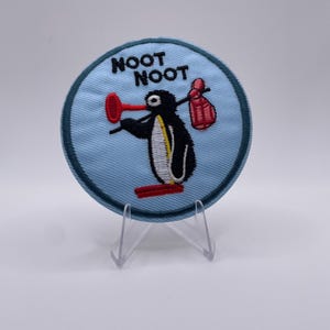 Op de afbeelding: Geborduurde patch met een cartoon pinguïn ontwerp. De pinguïn heeft een rode snavel en voeten, en houdt een rode ontstopper en een rode zak vast. De woorden "NOOT NOOT" zijn in zwart geborduurd op een lichtblauwe achtergrond.