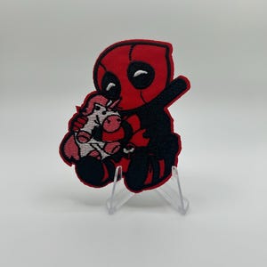Può includere: Una toppa ricamata raffigurante un personaggio in stile cartone animato con un costume rosso e nero, che tiene in mano un piccolo unicorno rosa e bianco. Il personaggio ha grandi occhi bianchi ed è seduto con l'elsa di una spada visibile dietro.