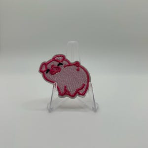 Könnte beinhalten: Ein hellrosa gestickter Aufnäher in Form eines kleinen Schweins. Das Schwein hat dunkelrosa gestickte Umrisse, schwarze gestickte Augen und eine kleine Schnauze. Der Aufnäher wird auf einem transparenten Kunststoffständer präsentiert.