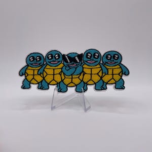 Puede incluir: Parche bordado con cinco personajes de tortugas de dibujos animados. Cuatro tortugas tienen los ojos abiertos, mientras que la tortuga central lleva gafas de sol. Las tortugas tienen cuerpos azules y patrones de caparazón amarillos. El parche se muestra sobre un soporte de plástico transparente sobre un fondo blanco.